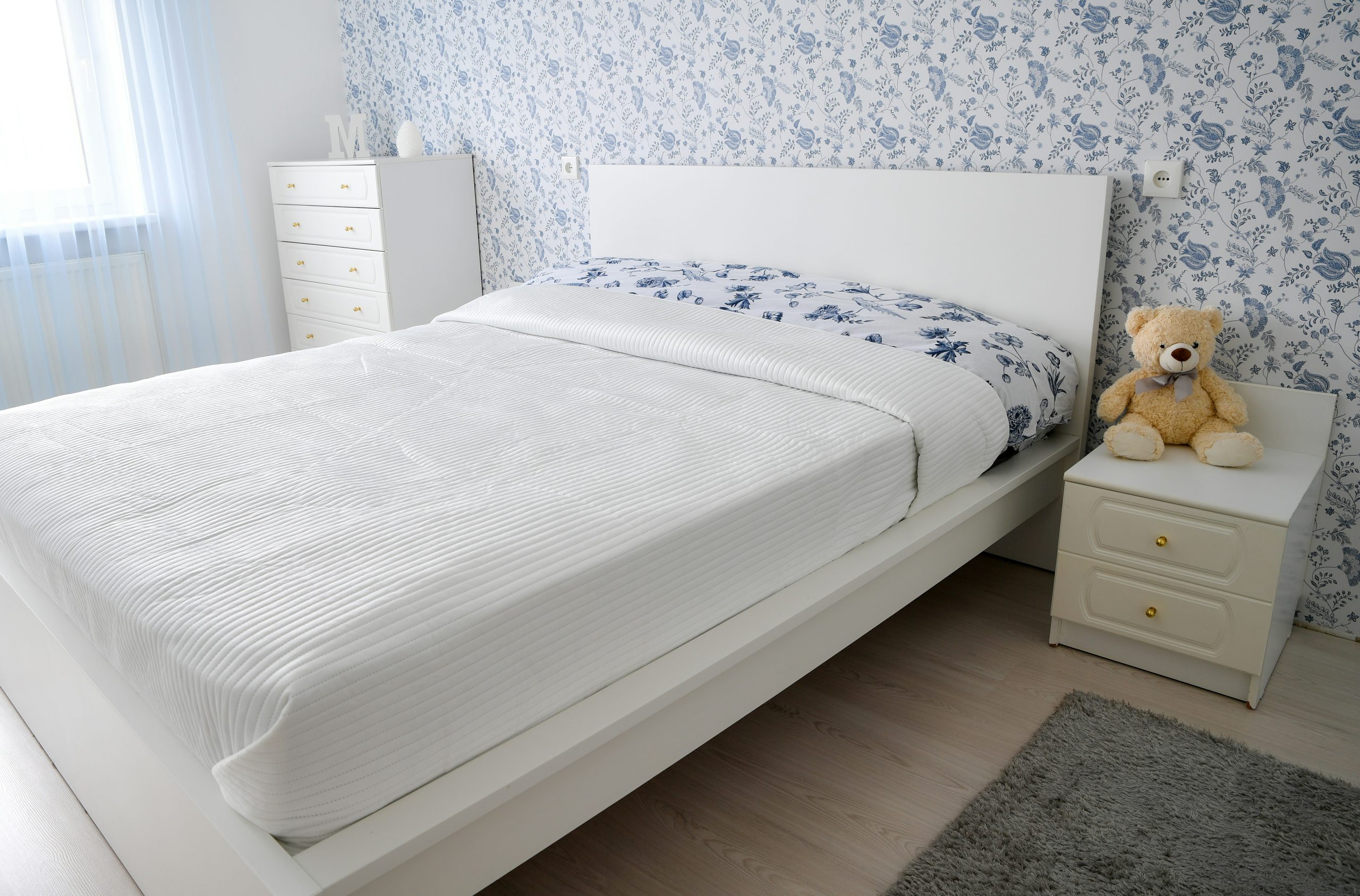 A bright and cozy modern bedroom interior featuring a white double bed, a matching nightstand with a teddy bear, and elegant blue and white floral wallpaper.📸 Photo by me IG: @margoevardson