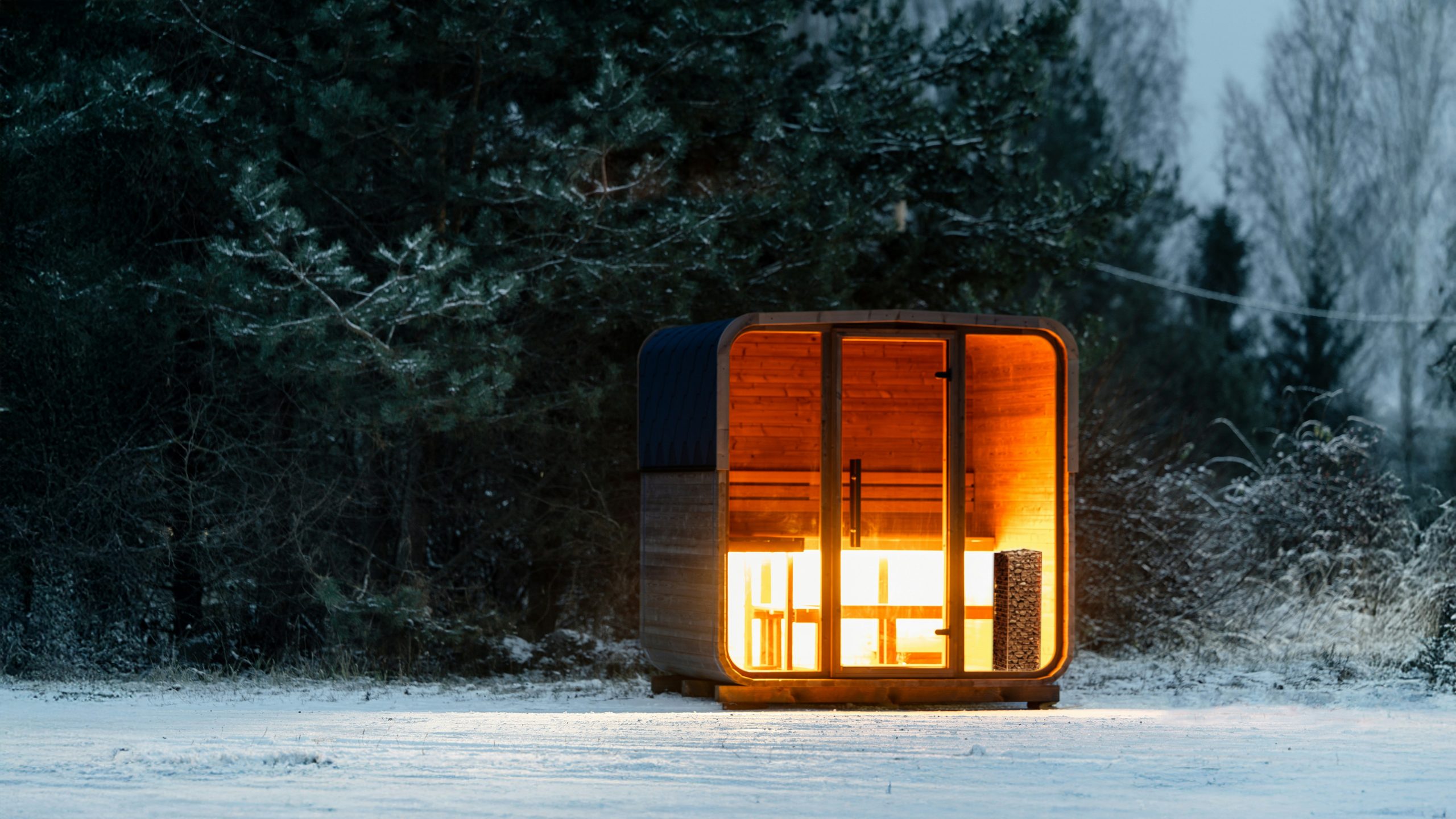 A cozy, illuminated wooden sauna cabin with a glass front, set in a snowy landscape surrounded by trees.