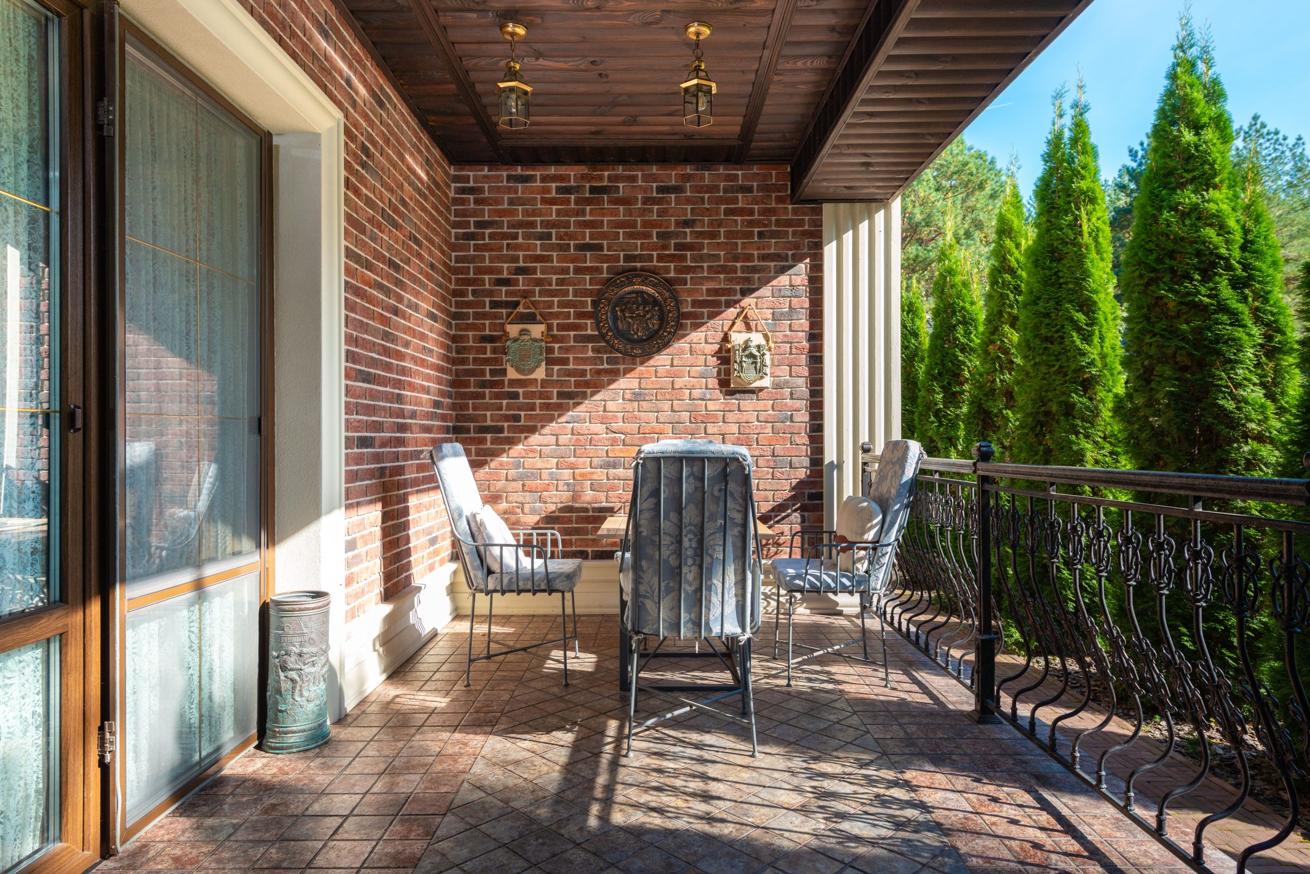A cozy terrace with chairs and a brick wall under a clear, sunny sky featuring lush green trees.