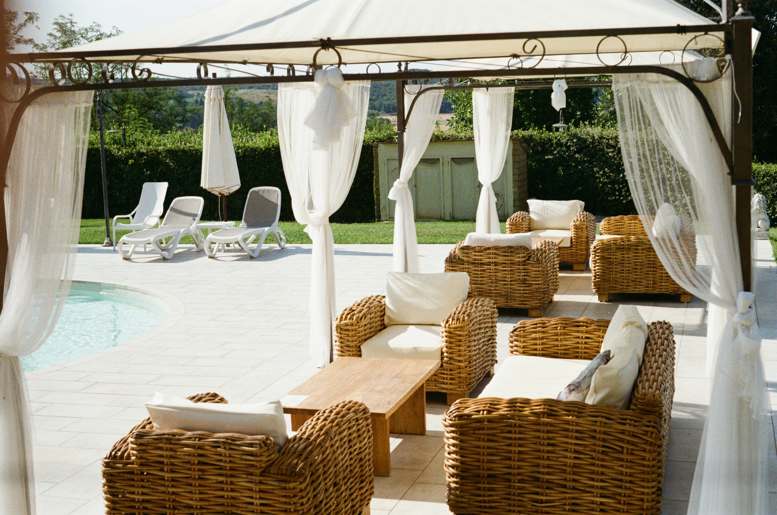 A draped pergola with wicker sofas overlooks a sunlit pool at an Italian countryside villa.