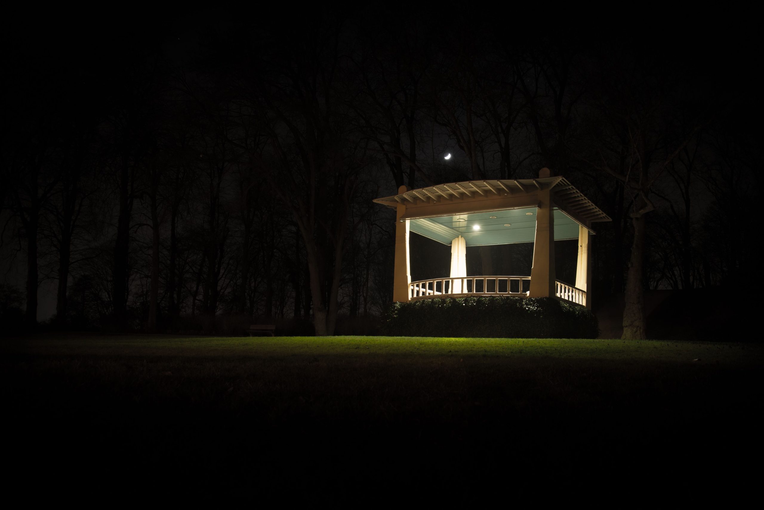 A serene nighttime scene featuring a well-lit gazebo surrounded by trees and a crescent moon.
