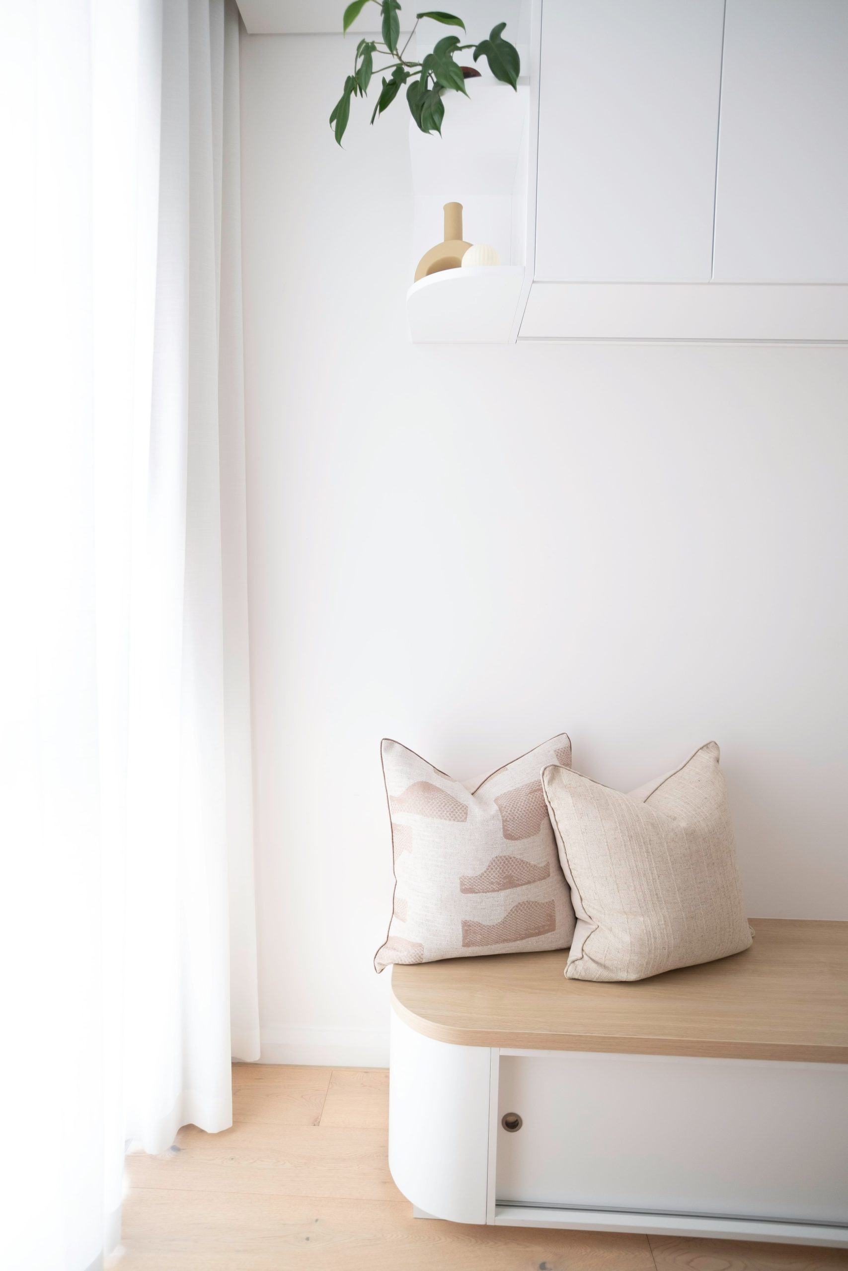 A soft, minimalist corner with two textured neutral cushions styled on a light timber bench. Sheer curtains filter gentle daylight into the space, creating a calm and airy interior. A simple sculptura