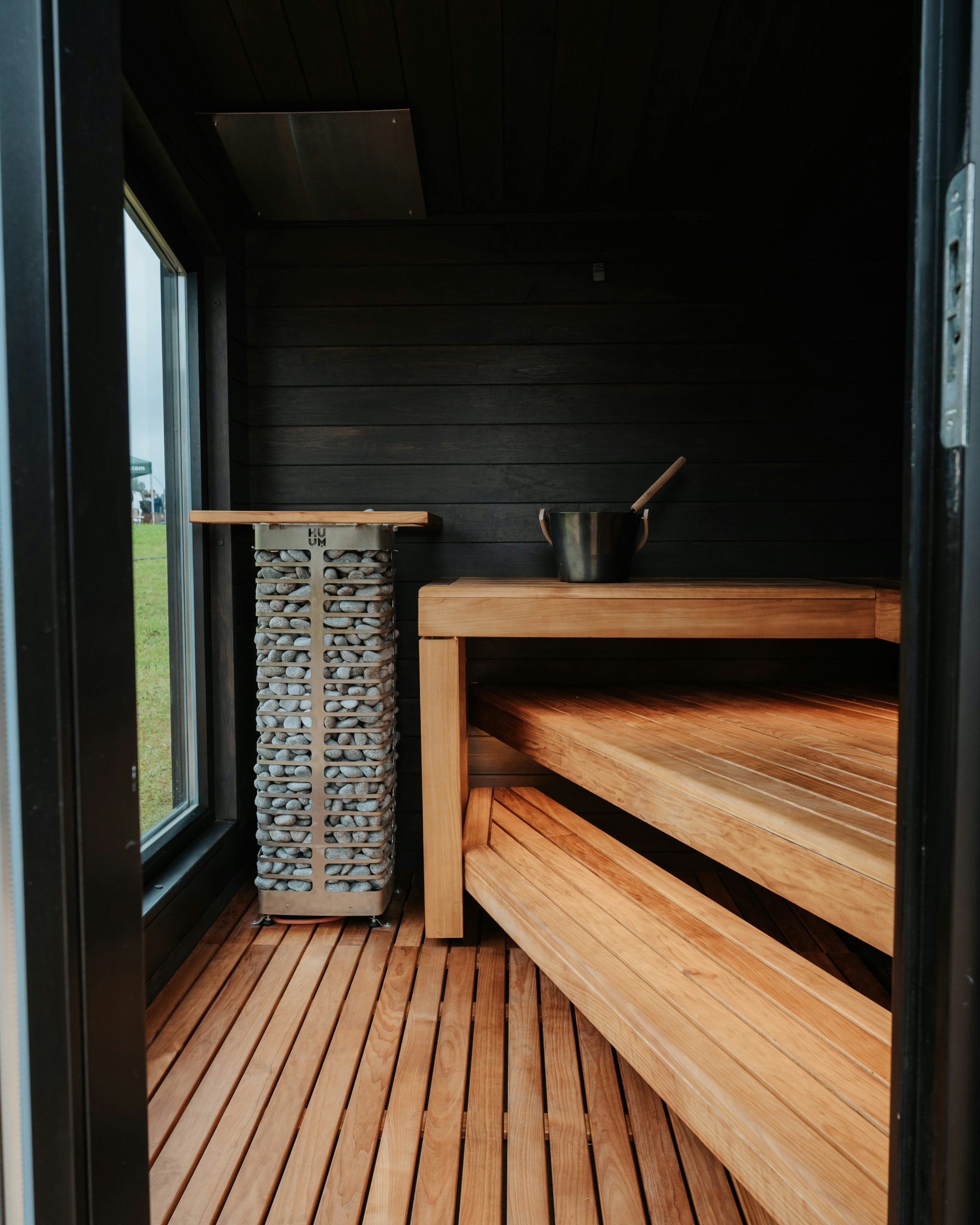 A stylish sauna interior featuring wooden benches, a tall stone heater, and contrasting dark wall panels. The design blends natural warmth with modern spa aesthetics for a calming wellness experience.