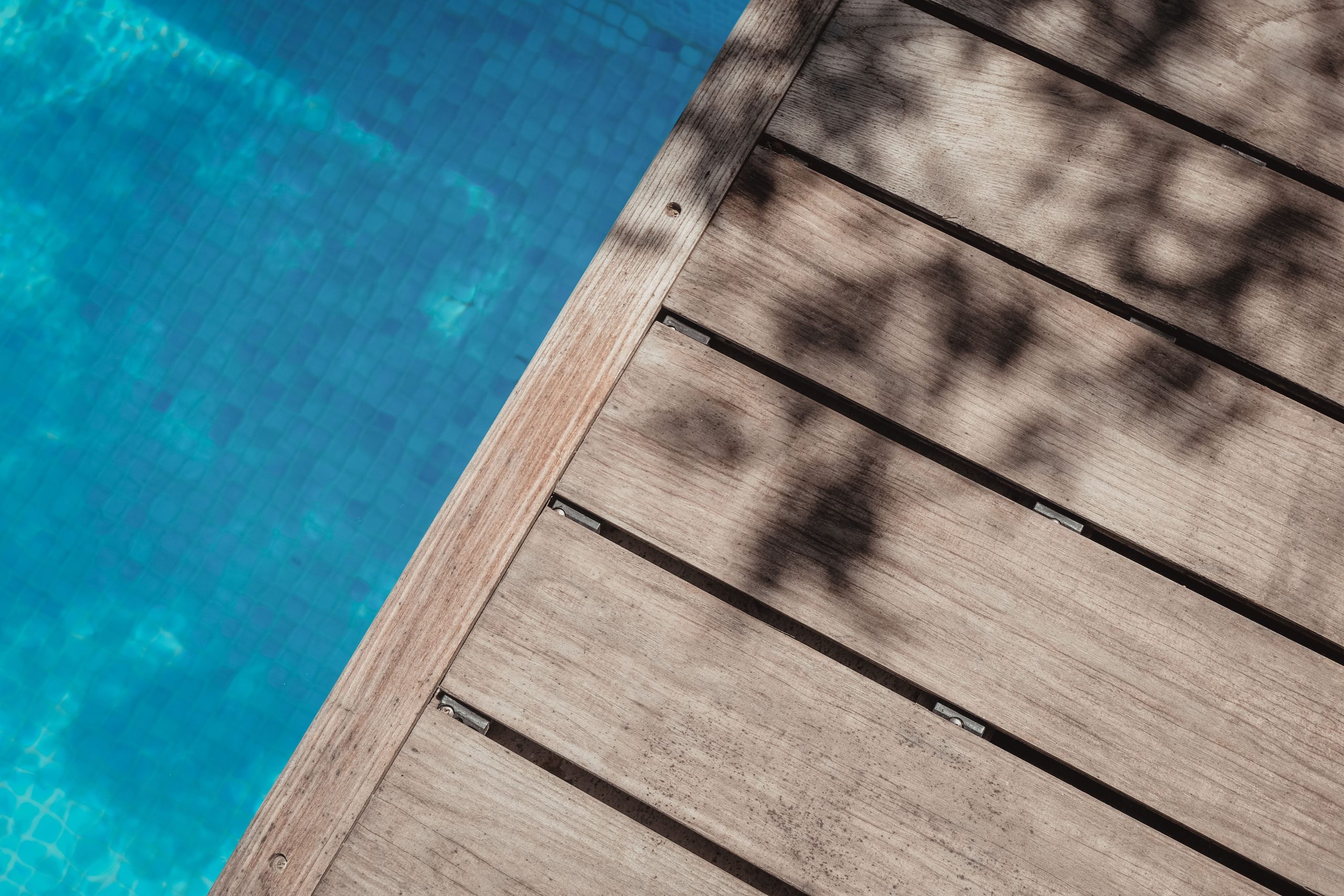 A wooden boardwalk casts shadows over a clear blue swimming pool, evoking a serene summer vibe.