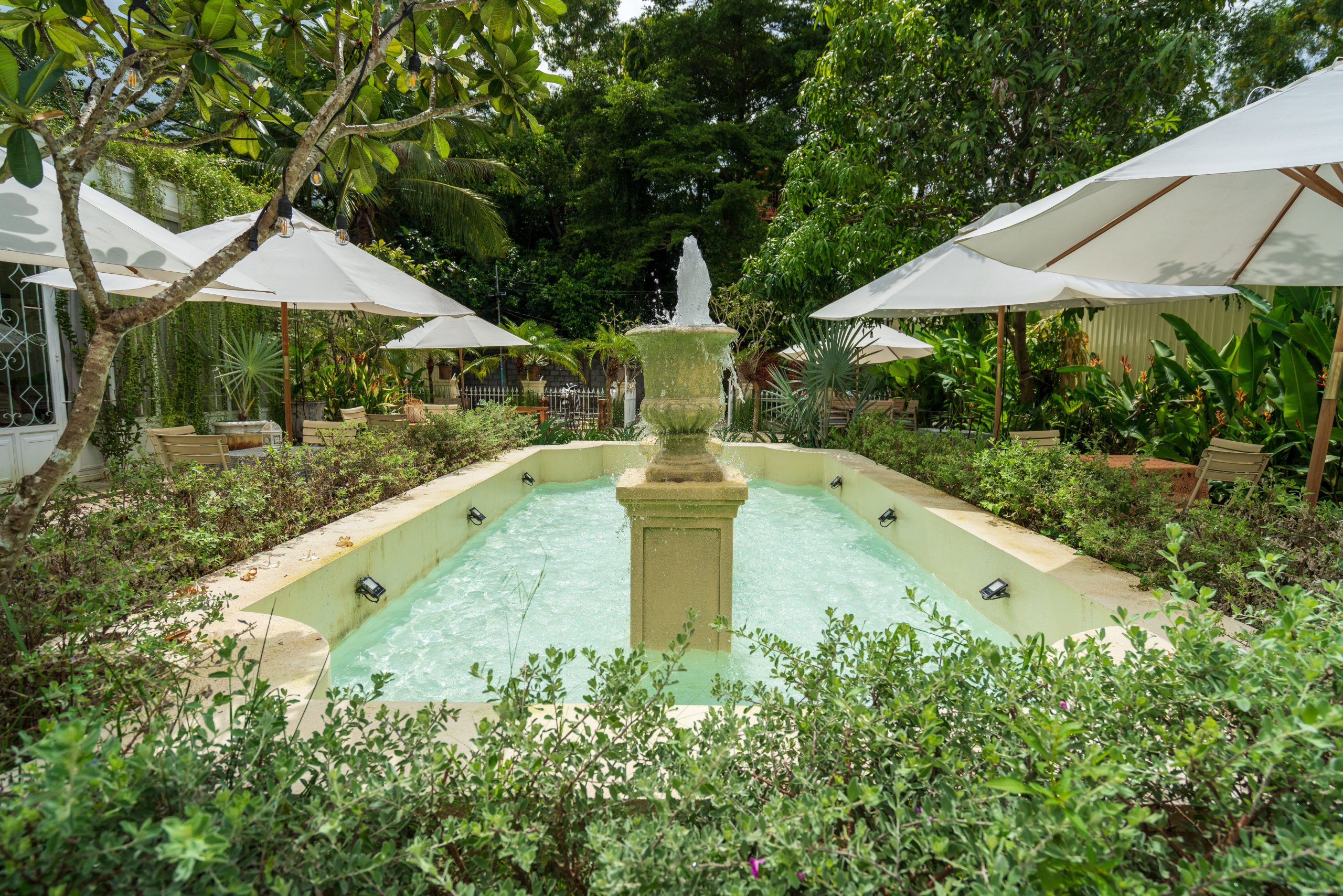 An elegant garden fountain surrounded by lush greenery, shaded by large umbrellas, creating a serene outdoor ambiance.