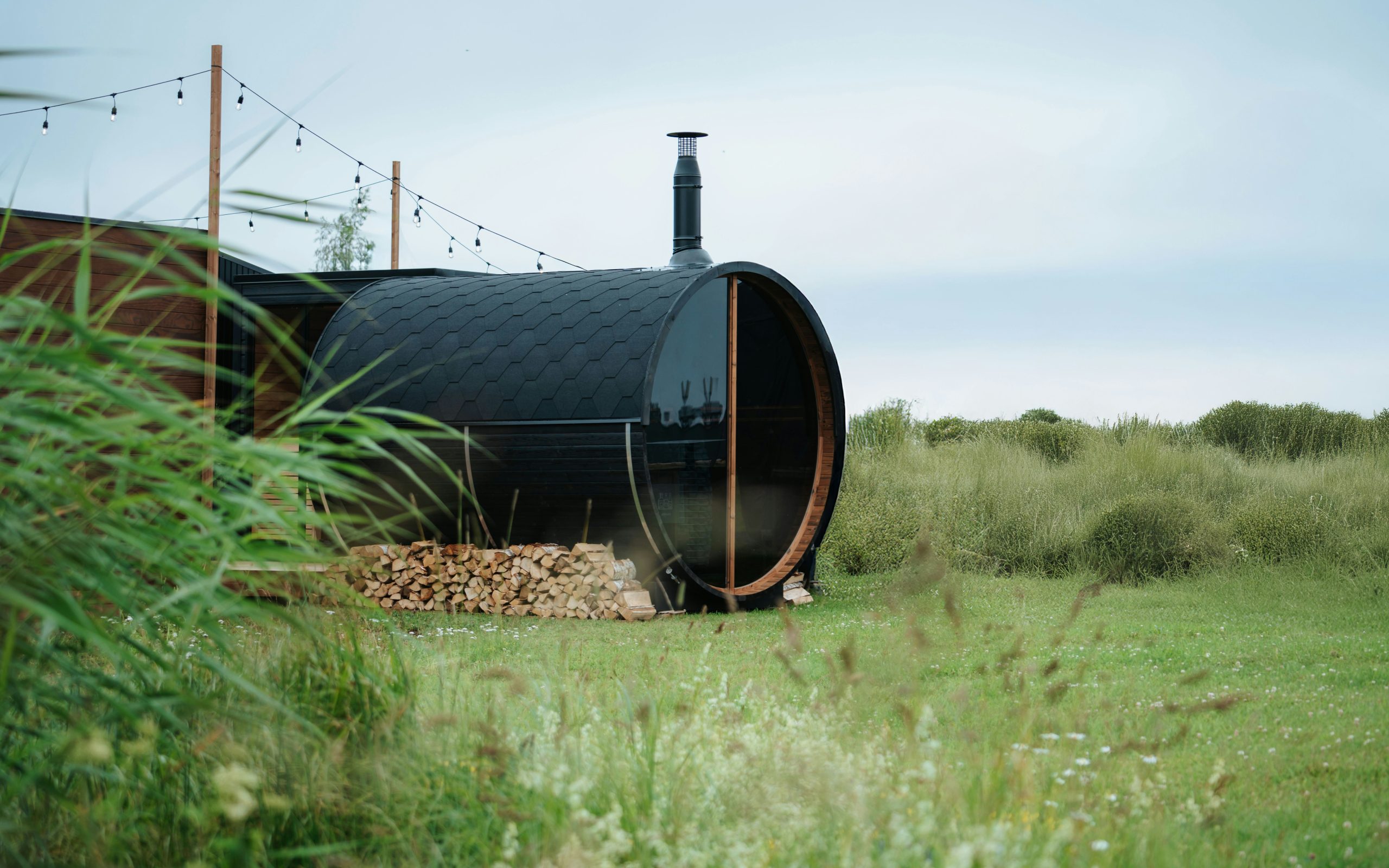 Auroom Halo black barrel sauna with black shingle roof and panoramic glass window, set in a natural green meadow with stacked firewood.