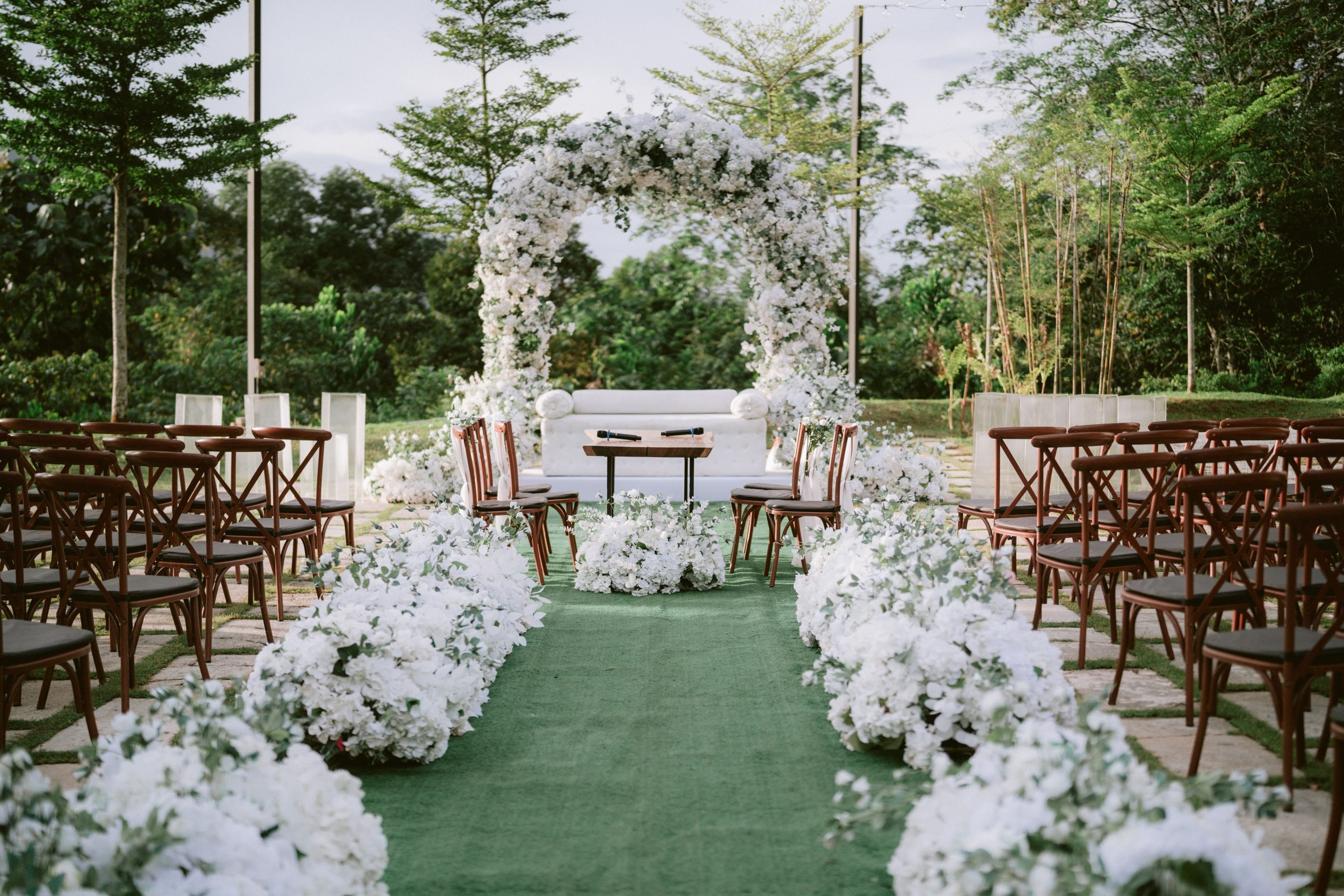 Beautiful outdoor wedding ceremony with white floral arch and chairs arranged beneath trees.