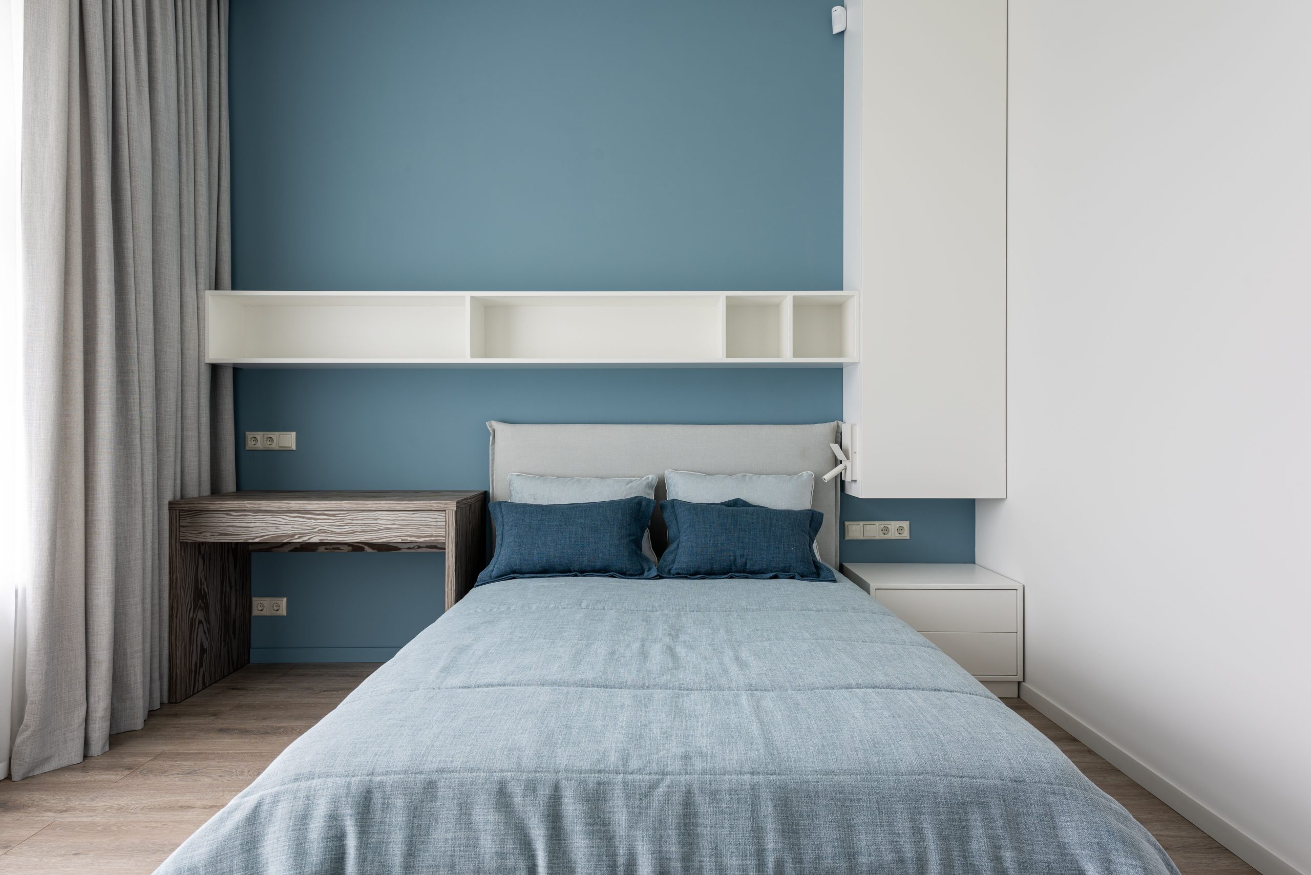 Contemporary simple interior of bedroom with white shelves and wooden table near window with curtains