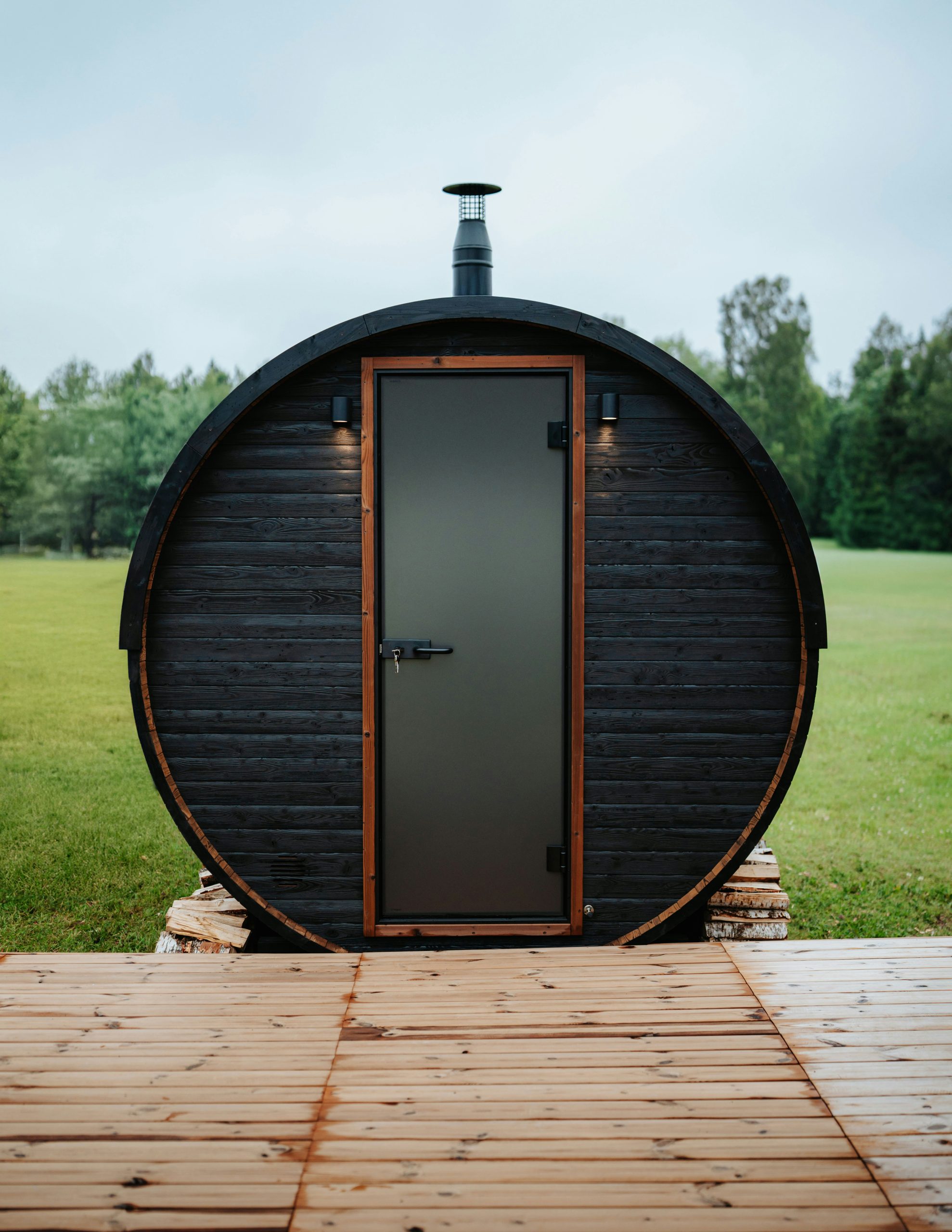 Front view of a round barrel sauna with a dark wood exterior and a modern door, set on a wooden deck in the outdoors.