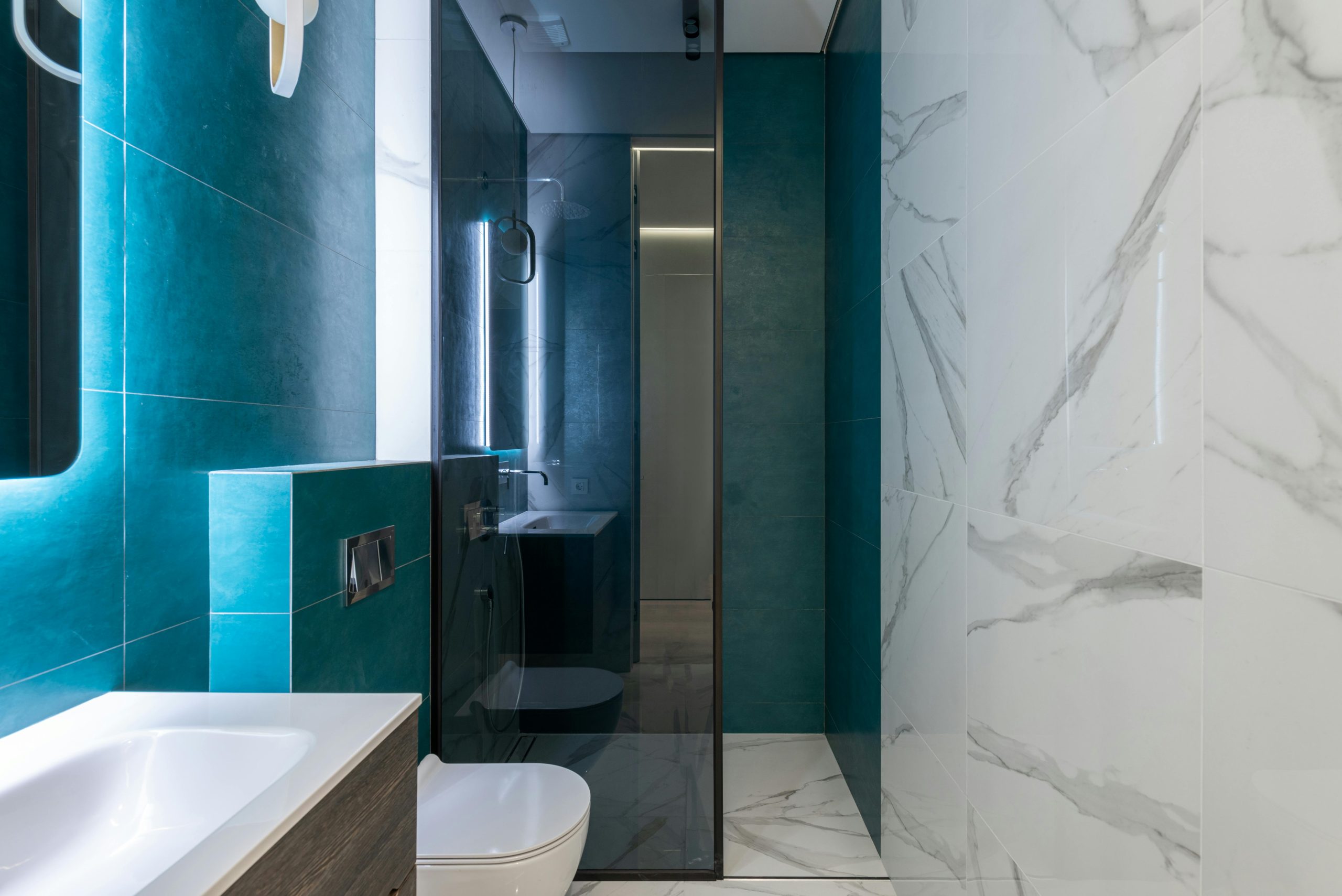 Glass shower cabin in contemporary bathroom with white ceramic toilet bowl and sink and tiled walls decorated with bright lights
