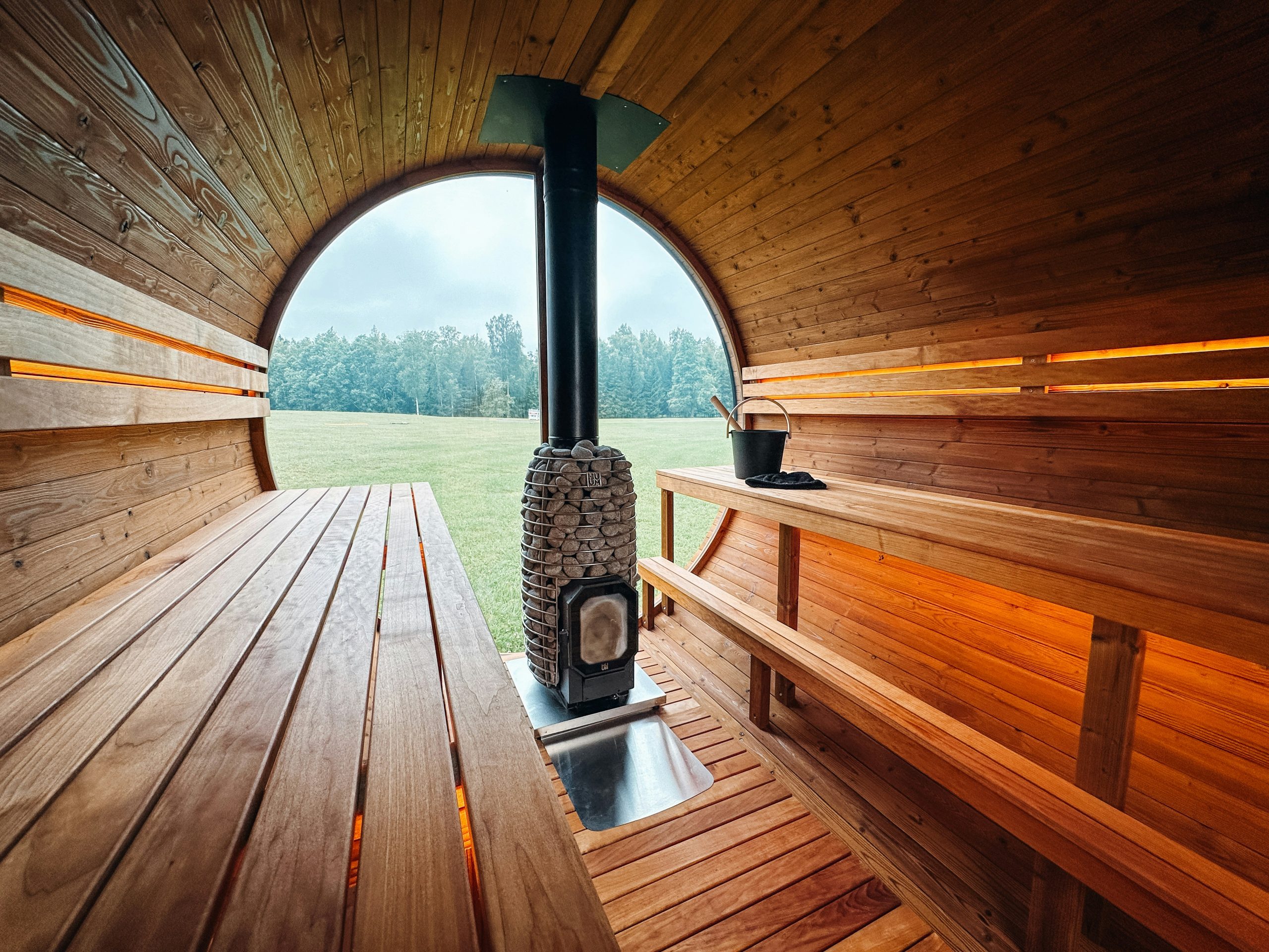 Inside a wooden sauna with wide benches and a stone-filled heater, opening to a panoramic glass window with a view of nature.