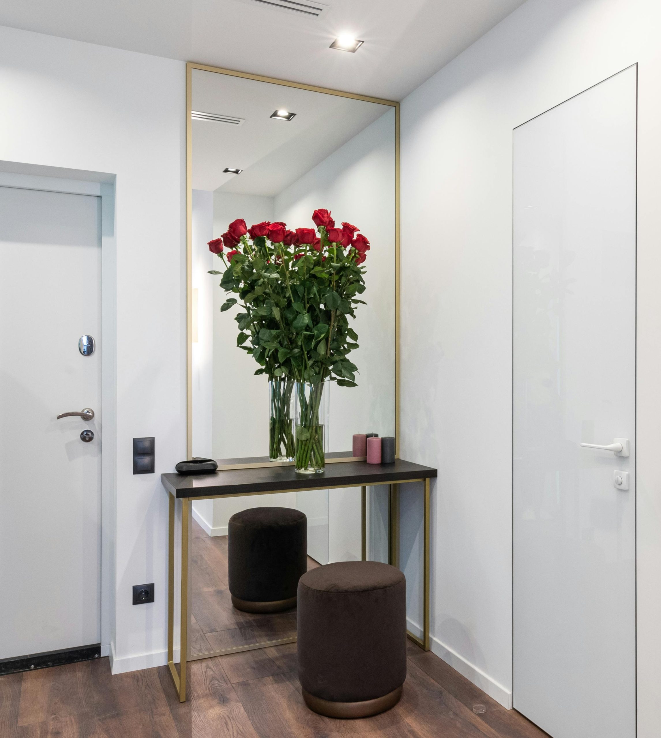 Interior of modern light apartment with mirror near table with bouquet of red roses in glass vase near pouf and doors