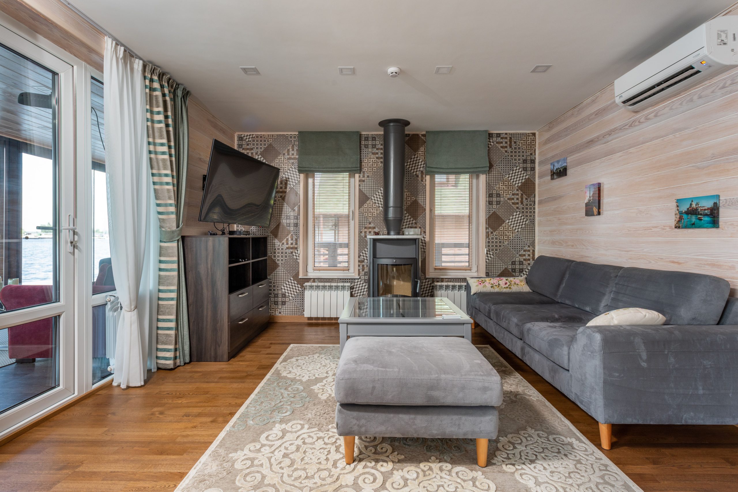 Modern design of country house with light wooden walls and carpet with ornament on parquet and dark gray furniture and curtains on wide balcony door and narrow windows
