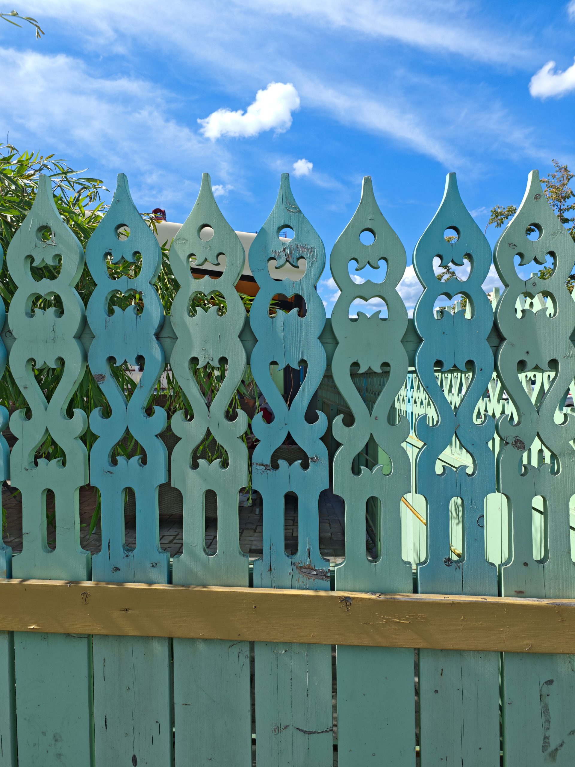 Ornate turquoise wooden fence with unique patterns against a vibrant blue sky.