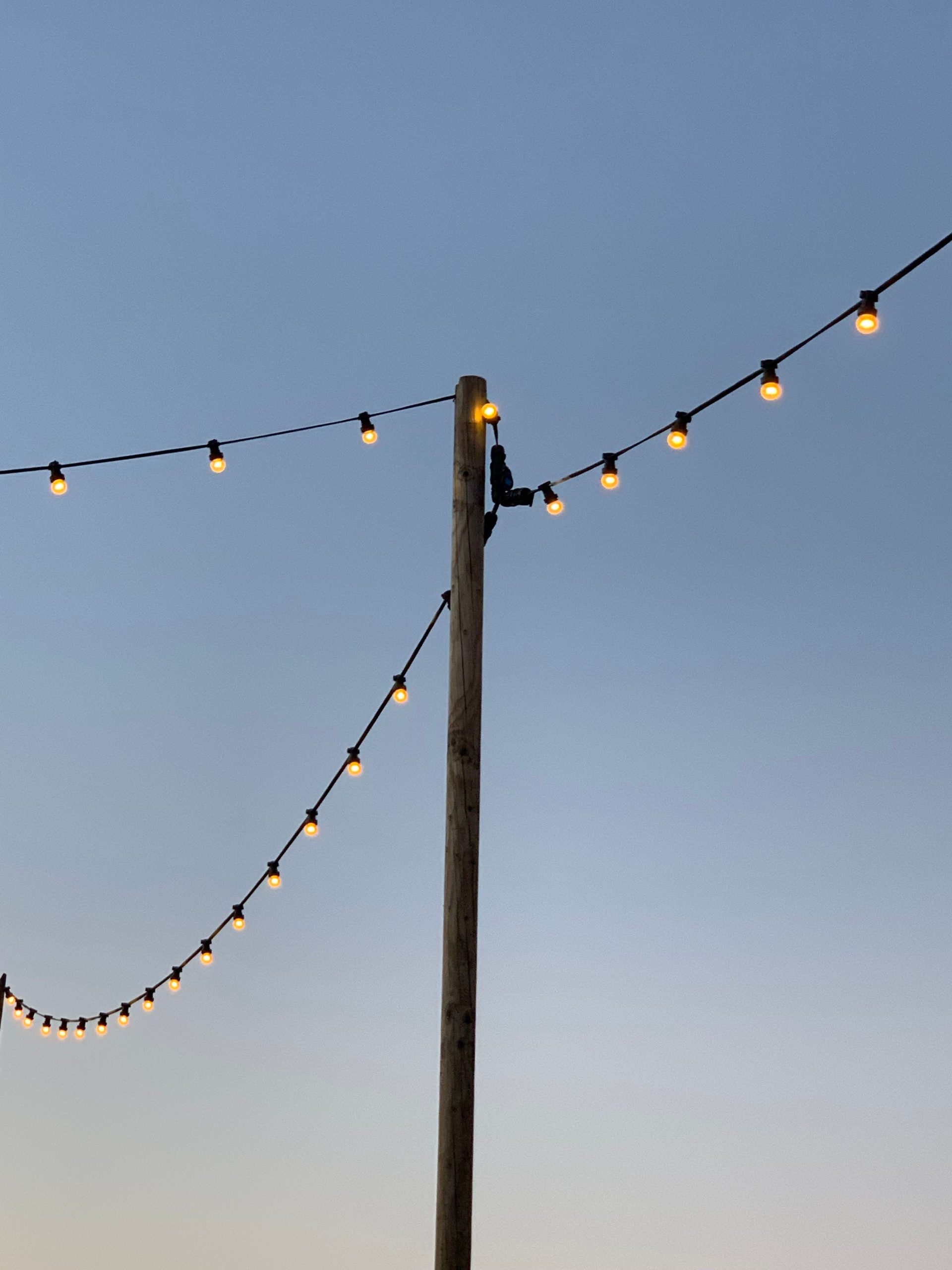 Warm glowing string lights hang against a clear blue sky, creating an inviting atmosphere.